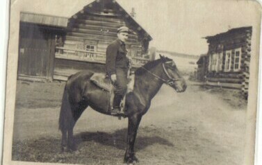 Młody mężczyzna siedzący na koniu, w tle drewniane zabudowania syberyjskiego kołchozu