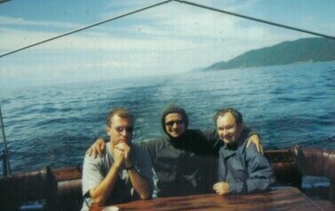 Trzech mężczyzn siedzących na pokładzie statku w części rufowej, w tle panorama jeziora bajkał