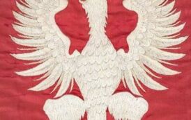 Na czerwonym tle wyhaftowany białą nicią stylizowany orzeł zwr&oacute;cony w prawo. Ponad jego głową znajduje się wyhaftowana złotą nicią korona